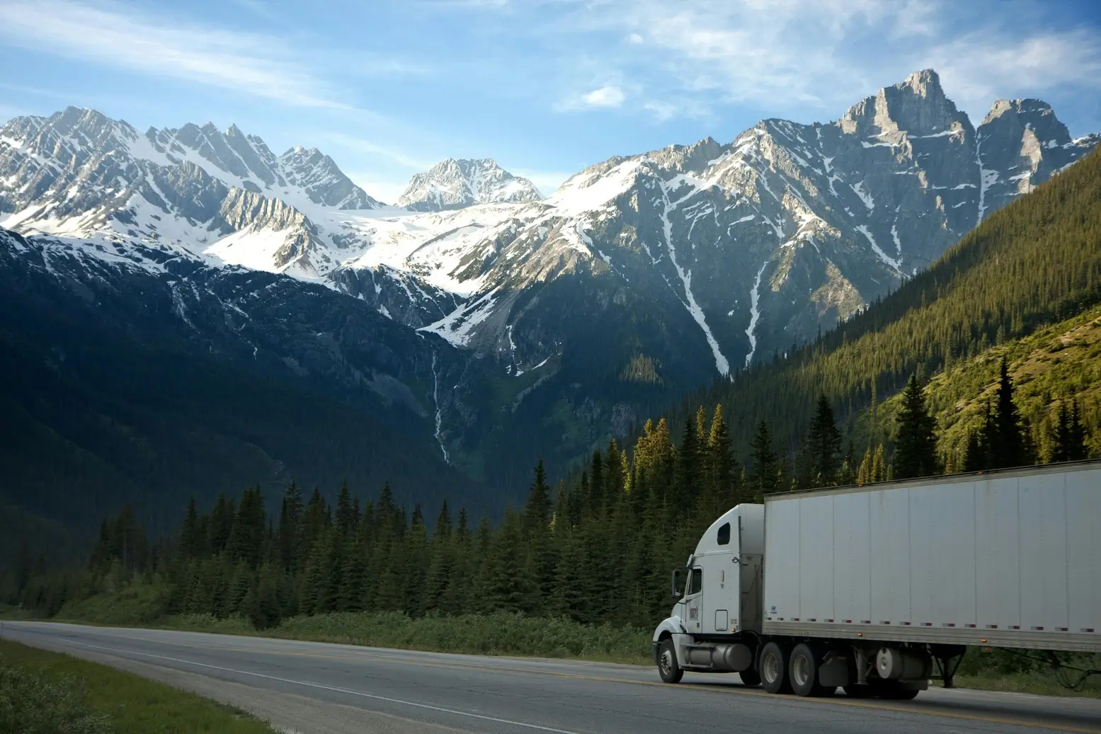 Snow-covered mountain range towering over a forest-lined highway with a semi truck driving through the valley